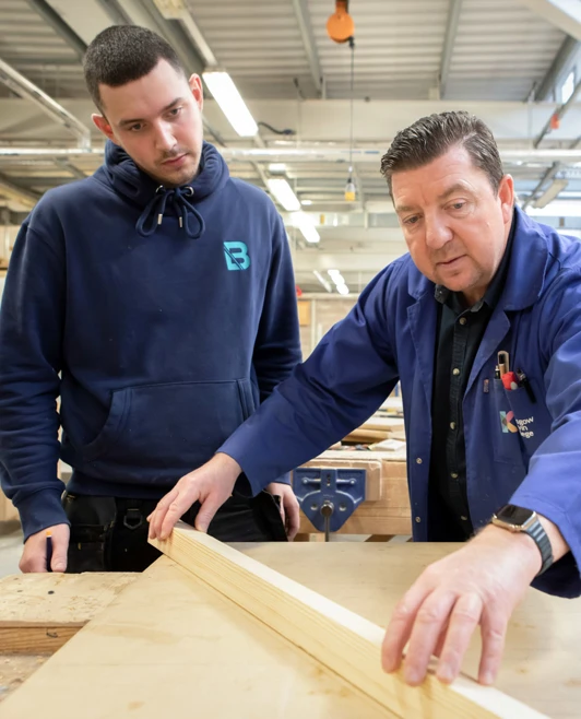 A joinery lecturer demonstrating woodworking techniques to a student, carefully inspecting a wooden plank on a workshop bench. A joinery lecturer demonstrating woodworking techniques to a student, carefully inspecting a wooden plank on a workshop bench.